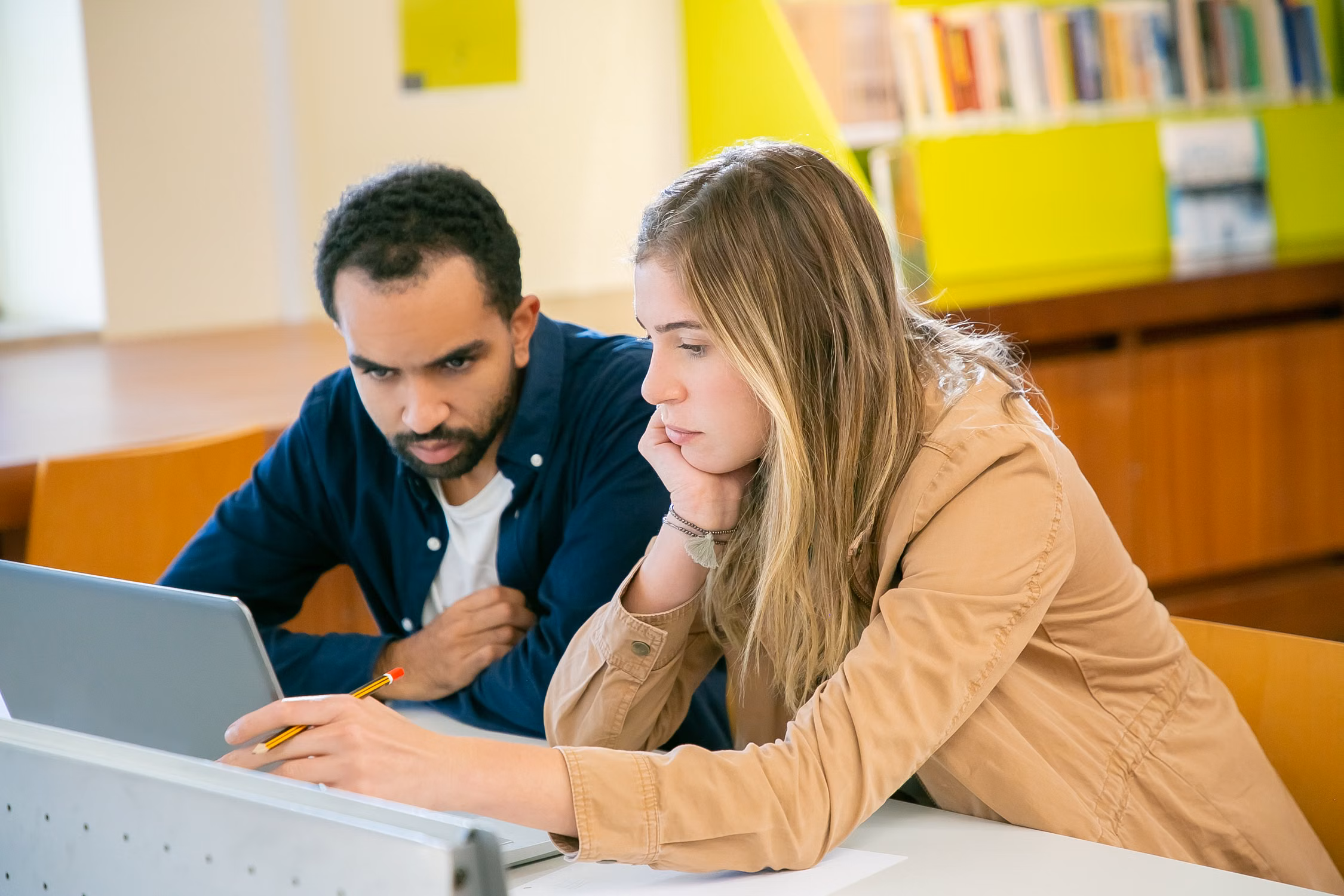 Two learners working together with a laptop and books in a library; illustrative stock photograph.